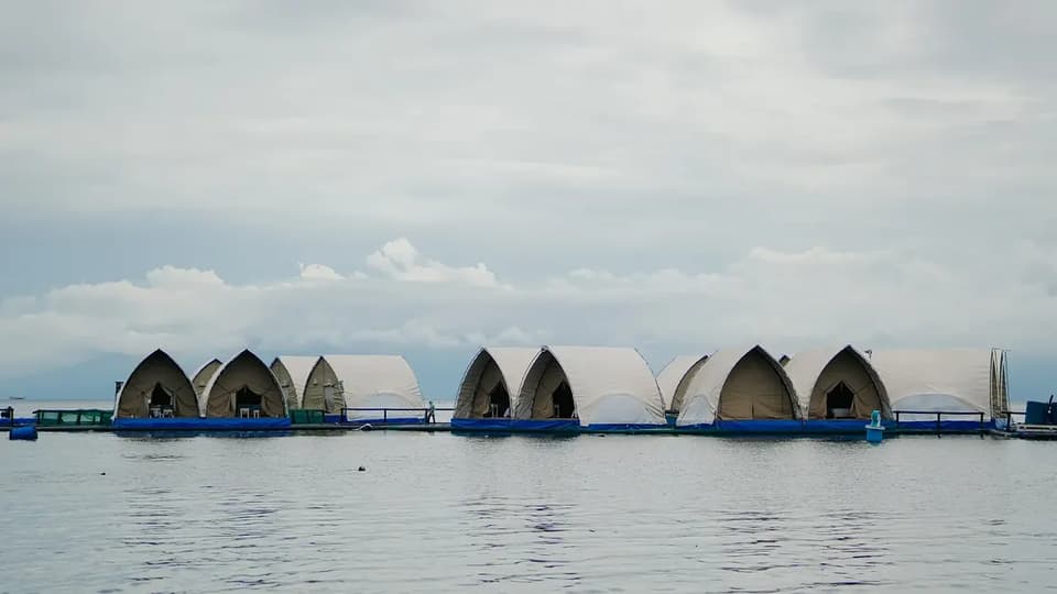 A row of peaked white glamphouses on blue pontoons at the Reef Resort, seen from the water under an overcast sky