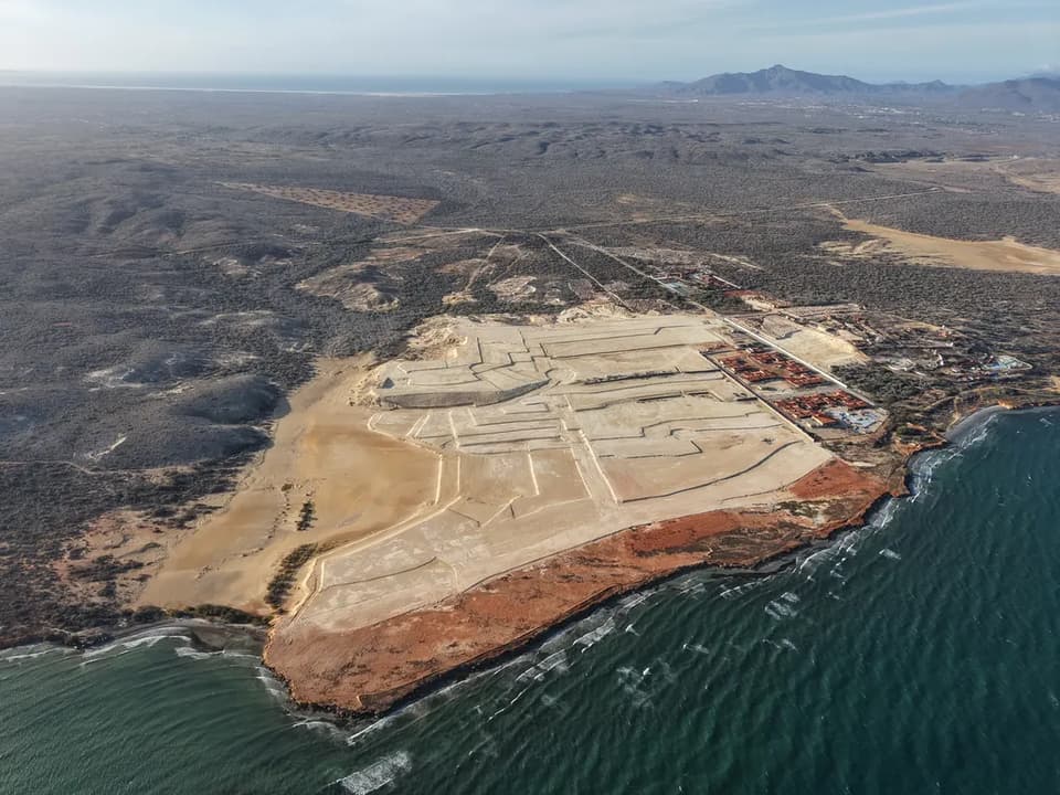 The CryptoCity site from the air at low sun: the graded headland cut into pale terraces, red cliffs along the shore and the dry hills of Margarita behind
