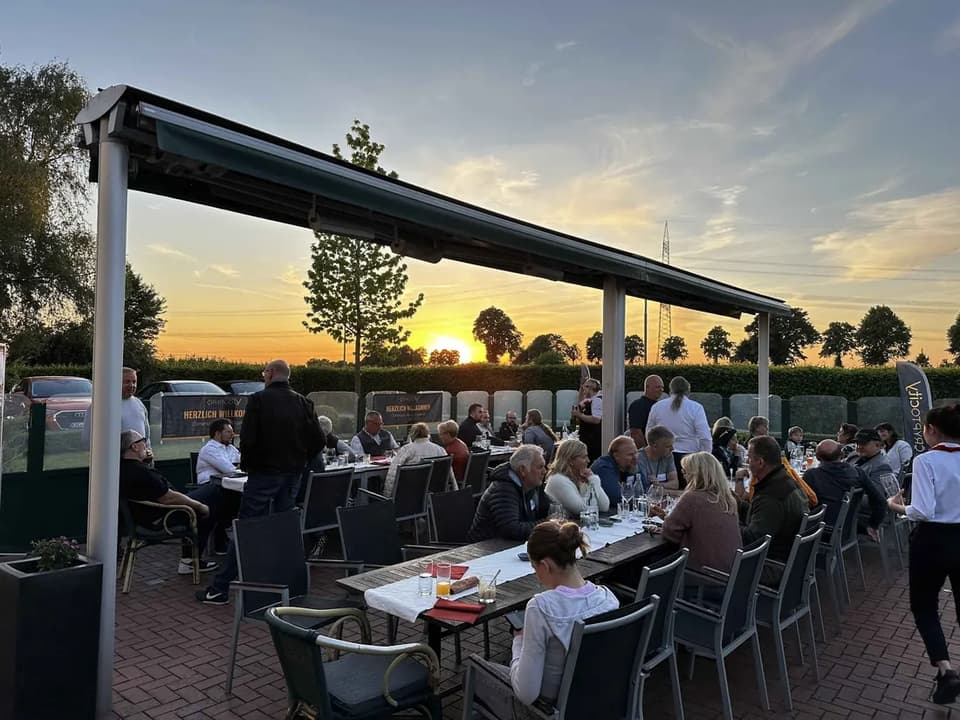 Members and prospective buyers at a long table under an awning at a CryptoCity gathering in Germany, the sun setting behind the hedge