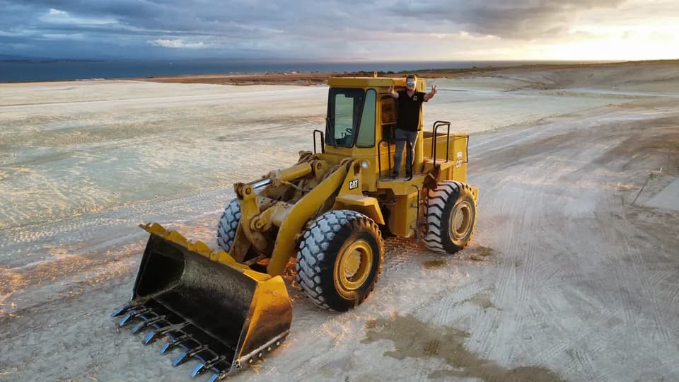 Tim Stern standing on the step of a wheel loader on the levelled site at dusk, the sea behind him