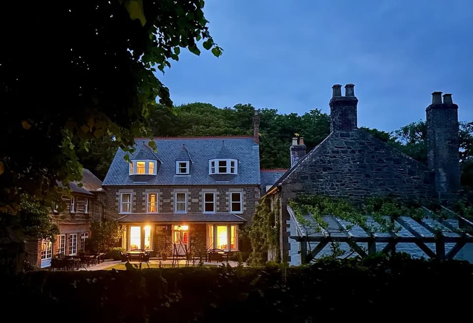 A granite country house glowing at dusk beneath Sark’s wooded hillside