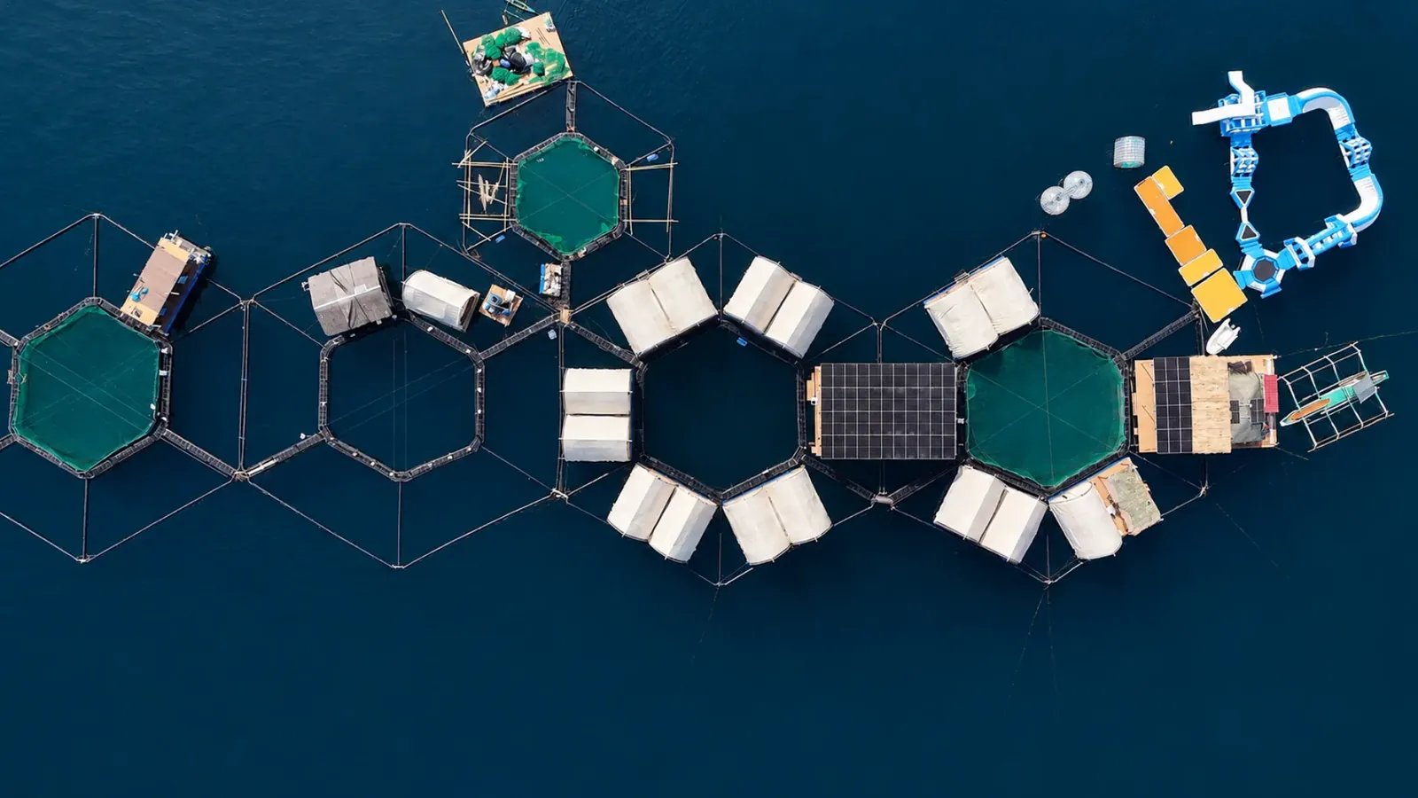 The Reef Resort from directly above: hexagonal fish pens with green netting, white glamphouses along the walkways, the solar array on the central deck and the inflatable water park moored alongside